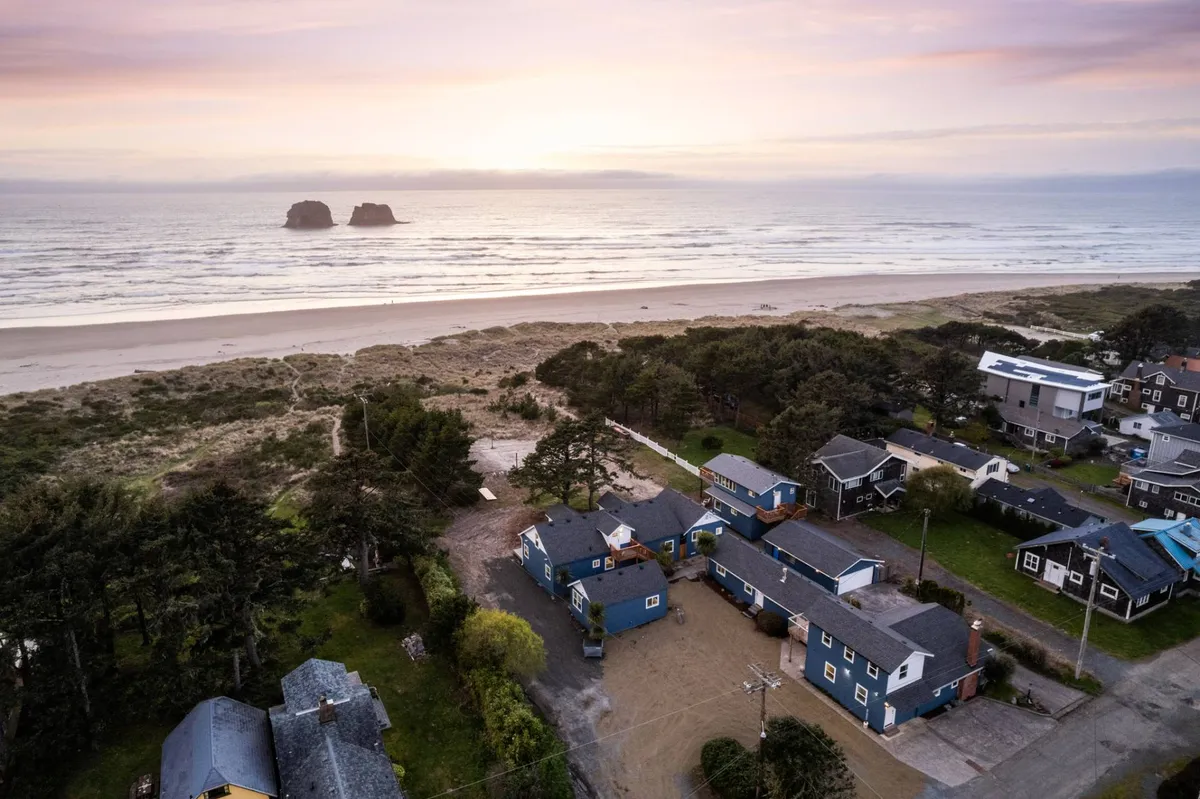 This image captures a wide view of the beach with gentle waves, soft sand, rock formations, and several cottages nestled among trees near the shore. The environment is peaceful, inviting families to enjoy beachcombing, building sandcastles, or relaxing together. The presence of both natural features and cottages suggests a direct connection between accommodations and the beach.
