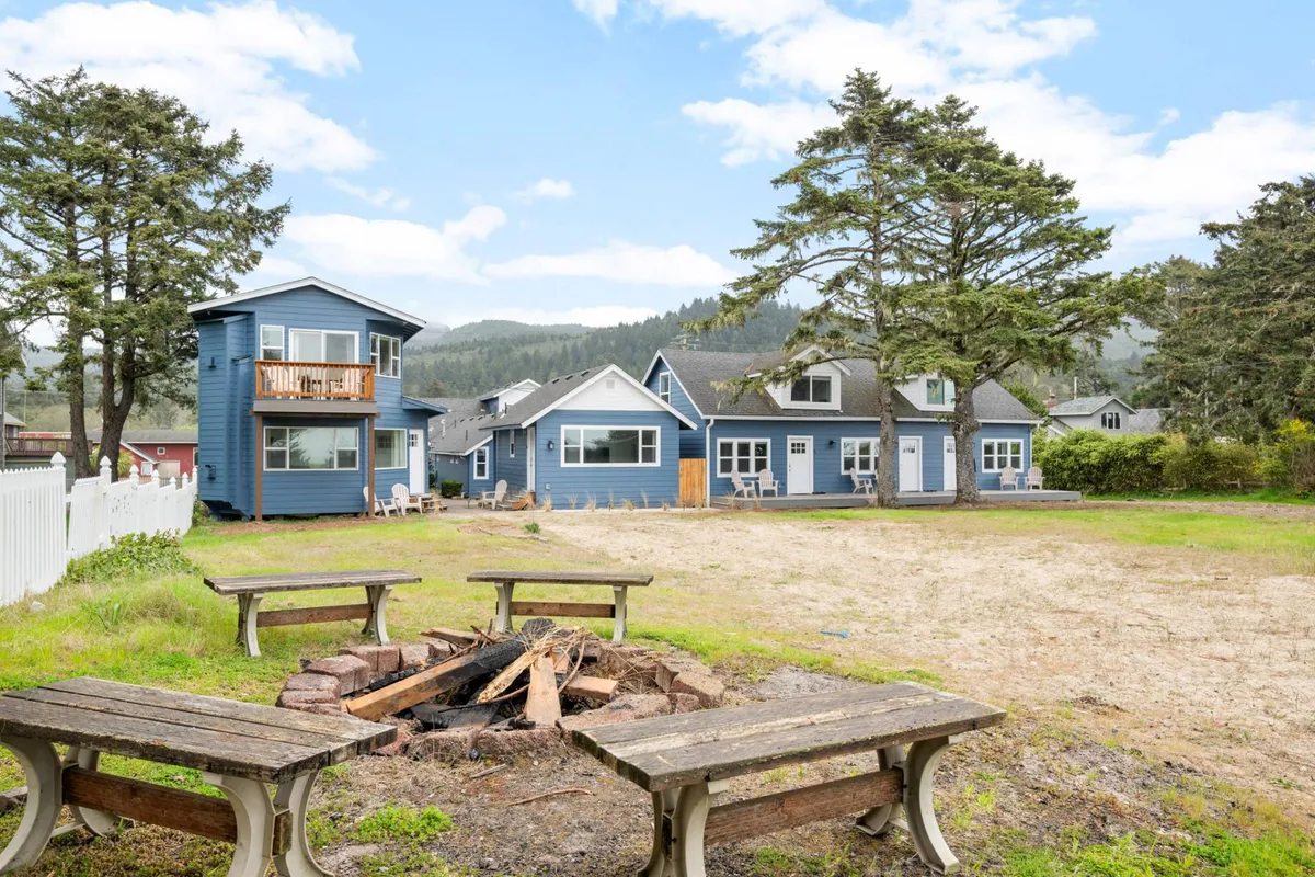 A cozy fire pit area surrounded by blue cottages and lush green trees. Several people are gathered around the fire, sharing stories and enjoying a relaxed atmosphere. The sunset casts a warm glow over the scene, emphasizing communal gathering and comfort.