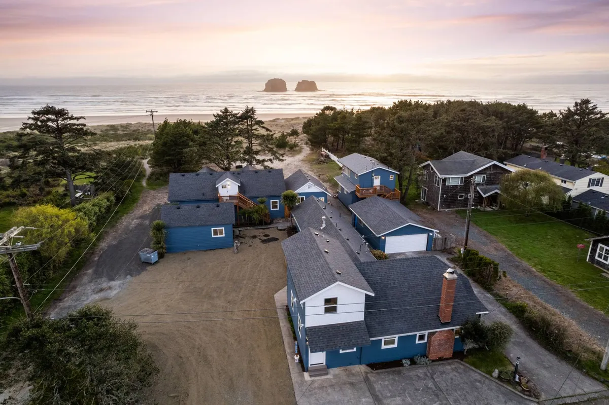 A sweeping exterior view featuring the ocean, dramatic rock formations, nearby houses, and trees. The scene evokes a sense of adventure—perfect for family beach walks, tide pool exploration, and gathering around a fire pit after a day of fun.