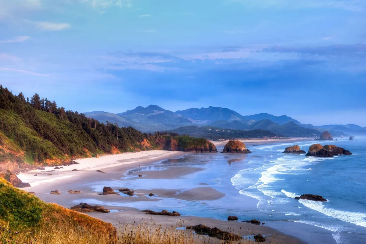A breathtaking panoramic view of the coastline, with sandy beach, ocean waves, and distant cliffs and mountains. The scene invites thoughts of beachcombing, volleyball on the sand, and exploring the natural beauty just beyond the inn.