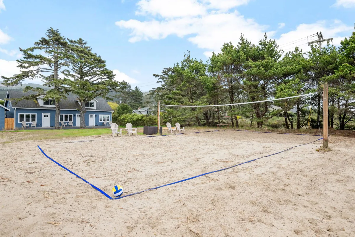The image captures a sandy volleyball court bordered by a volleyball net, with inviting lawn chairs scattered nearby. Mature trees frame the open space and the mood is playful and relaxed, ideal for friendly competitions or cheering on a lively game below from a nearby deck.