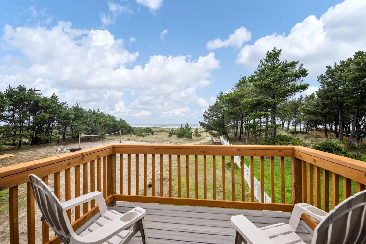 A private deck with a wooden railing overlooks sweeping ocean views framed by trees. Two comfortable chairs are positioned to enjoy the panorama, inviting guests to relax, read, or socialize while surrounded by nature. The sky hints at sunset, adding warmth to the tranquil setting.