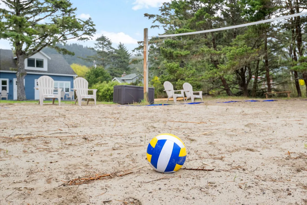 An outdoor sand volleyball court framed by surrounding trees, with a group actively playing volleyball. Nearby, guests lounge in chairs, chatting and cheering, while others relax in the inviting outdoor seating area. The space exudes playful energy and encourages group interaction.