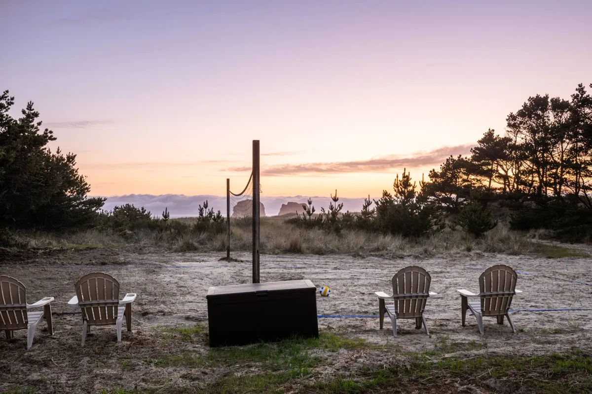 A serene, wide-angle view over a volleyball net and sandy court with Adirondack chairs in the foreground. The background features distant rock formations, possibly Twin Rocks, and a colorful sunset sky, combining the thrill of activity with the tranquil natural beauty of the coast.