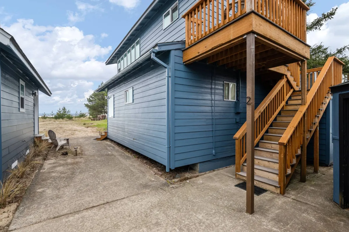 The image features a welcoming outdoor seating area on a wooden deck with blue siding, accompanied by stairs leading down to the beach. The setup suggests an ideal spot for morning coffee or sharing a sunset toast, with an atmosphere perfect for intimate moments.